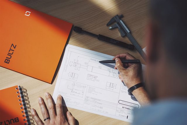 Mesa de trabajo con plano técnico de un resorte de gas Stabilus, calibre y cuaderno de Bultz